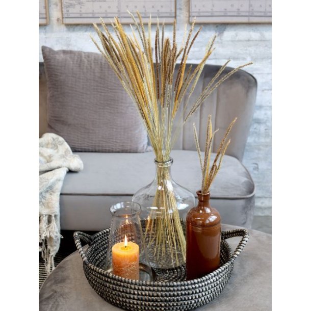 Den mocca-brune keramikflaske st�r i en flettet bakke p� en puf sammen med en stor glaskaraffel med t�rrede str� og en t�ndt orange bloklys i en glasholder, foran en gr� sofa.