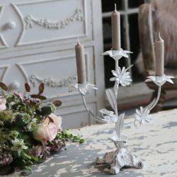 En hvid trearmet lysestage i fransk landstil, dekoreret med fine metalblomster og blade. Lysestagen har et slidt, patineret udtryk og er sat med tre t�ndte, st�vede brune kronelys. Den st�r p� et rustikt, hvidmalet tr�bord med afskallet maling ved siden af en smuk blomsterbuket med lyser�de roser. I den sl�rede baggrund ses en elegant, hvid kommode med krummelurer, der fuldender den romantiske og franske stemning.