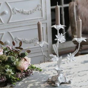 En hvid trearmet lysestage i fransk landstil, dekoreret med fine metalblomster og blade. Lysestagen har et slidt, patineret udtryk og er sat med tre t�ndte, st�vede brune kronelys. Den st�r p� et rustikt, hvidmalet tr�bord med afskallet maling ved siden af en smuk blomsterbuket med lyser�de roser. I den sl�rede baggrund ses en elegant, hvid kommode med krummelurer, der fuldender den romantiske og franske stemning.