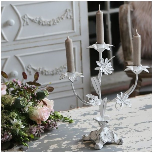 En hvid trearmet lysestage i fransk landstil, dekoreret med fine metalblomster og blade. Lysestagen har et slidt, patineret udtryk og er sat med tre t�ndte, st�vede brune kronelys. Den st�r p� et rustikt, hvidmalet tr�bord med afskallet maling ved siden af en smuk blomsterbuket med lyser�de roser. I den sl�rede baggrund ses en elegant, hvid kommode med krummelurer, der fuldender den romantiske og franske stemning.