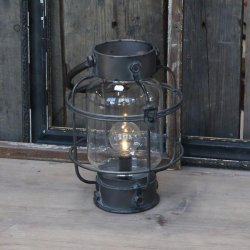 En rustik rund staldlanterne i antik sort metal placeret p� et r�t tr�bord. Lanternen er t�ndt og spreder et varmt lys gennem det klare glas.