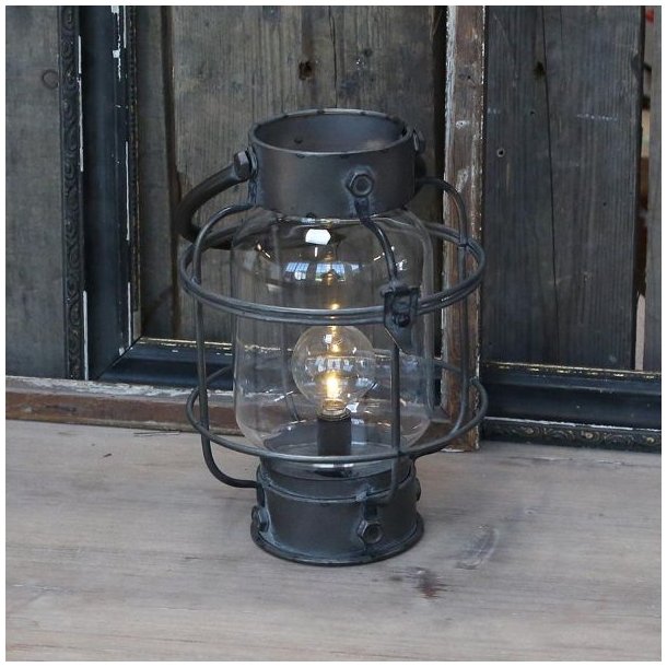 En rustik rund staldlanterne i antik sort metal placeret p� et r�t tr�bord. Lanternen er t�ndt og spreder et varmt lys gennem det klare glas.