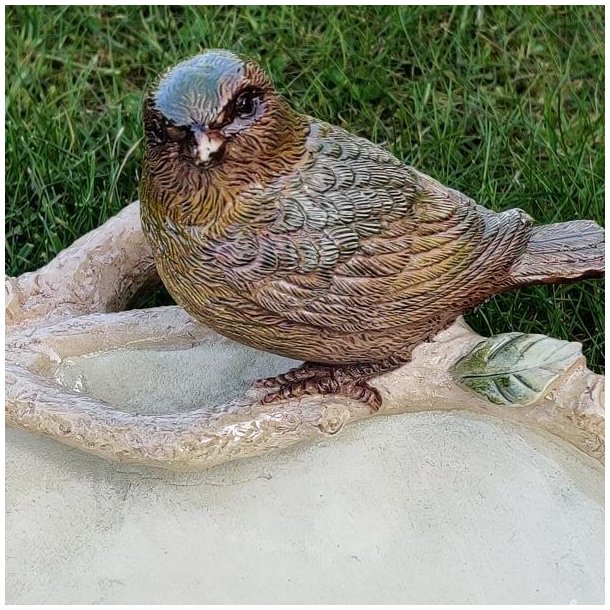 Fokus p� den anden fuglefigur p� kanten af fuglebadet med synlige detaljer i fjerdragten og de gr�nne blade.