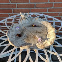 Bladformet fuglebad med vand og blomster p� et hvidt havebord med en murstensv�g i baggrunden.