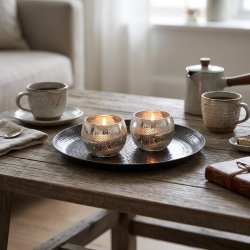 Stemningsfuldt 1:1 milj�billede af to sm� runde fyrfadsstager i antik s�lv med graveret bladm�nster og t�ndte lys, st�ende p� et tray-fad p� et veld�kket kaffebord.