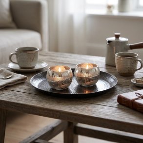 Stemningsfuldt 1:1 milj�billede af to sm� runde fyrfadsstager i antik s�lv med graveret bladm�nster og t�ndte lys, st�ende p� et tray-fad p� et veld�kket kaffebord.