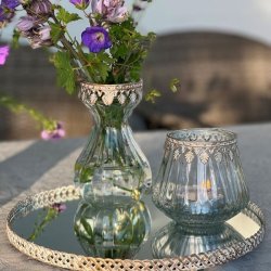 Et stemningsfuldt milj�billede i 1:1 format, der viser den t�ndte glasfyrfadsstage med antik guld dekorationskant p� en spejlbakke. Ved siden af st�r en matchende glasvase med en buket lilla storken�b, der reflekteres i spejloverfladen mod en hyggelig, sl�ret havebaggrund.