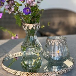 Et stemningsfuldt milj�billede i 1:1 format, der viser den t�ndte glasfyrfadsstage med antik guld dekorationskant p� en spejlbakke. Ved siden af st�r en matchende glasvase med en buket lilla storken�b, der reflekteres i spejloverfladen mod en hyggelig, sl�ret havebaggrund.