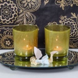 Et stemningsfuldt milj�billede i 1:1 format, der viser to t�ndte fyrfadsstager i klart glas med det specifikke gr�nne bladm�nster, baseret p� image_86.png. De st�r p� en rund spejlbakke ved siden af en lille dekorativ alf-figur, mod en baggrund med et matchende sort og guld m�nstret tapet.