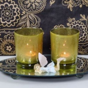 Et stemningsfuldt milj�billede i 1:1 format, der viser to t�ndte fyrfadsstager i klart glas med det specifikke gr�nne bladm�nster, baseret p� image_86.png. De st�r p� en rund spejlbakke ved siden af en lille dekorativ alf-figur, mod en baggrund med et matchende sort og guld m�nstret tapet.