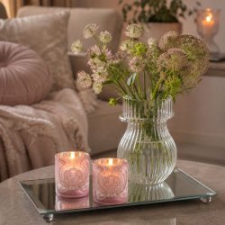 To mat rosa fyrfadsstager med velourm�nster og t�ndte lys st�ende p� en rektangul�r spejlbakke ved siden af en glasvase med blomster.