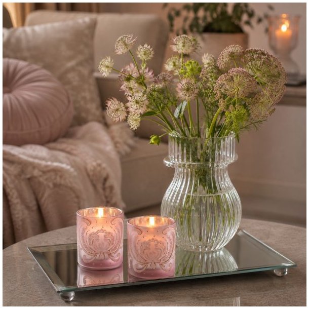 To mat rosa fyrfadsstager med velourm�nster og t�ndte lys st�ende p� en rektangul�r spejlbakke ved siden af en glasvase med blomster.
