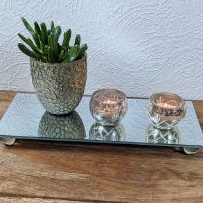 To m�nstrede fyrfadsstager i antique s�lv med t�ndte lys og en gr�n potteplante arrangeret p� et rektangul�rt spejlplateau.