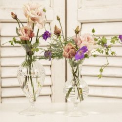 To elegante, facetslebne glasvaser p� fod, der st�r p� et hvidt bord foran en hvid jalousil�ge. Begge vaser er fyldt med vand og lette, romantiske blomster i sarte lyser�de og violette nuancer samt fine, gr�nne grene, der skaber en luftig og sommerlig stemning.