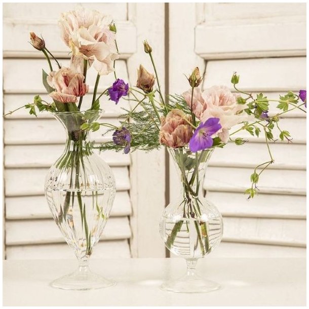 To elegante, facetslebne glasvaser p� fod, der st�r p� et hvidt bord foran en hvid jalousil�ge. Begge vaser er fyldt med vand og lette, romantiske blomster i sarte lyser�de og violette nuancer samt fine, gr�nne grene, der skaber en luftig og sommerlig stemning.