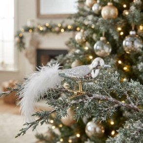 S�lvfarvet pyntefugl i glas med fjerhale sat fast p� en grangren p� et juletr� med hyggelige julelys i baggrunden.