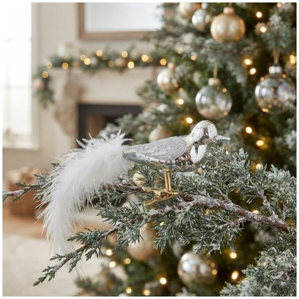 S�lvfarvet pyntefugl i glas med fjerhale sat fast p� en grangren p� et juletr� med hyggelige julelys i baggrunden.