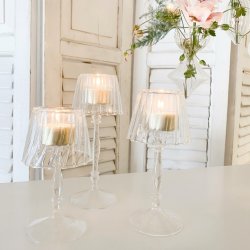 Tre elegante glasfyrfadsstager p� h�je stilke, formet som sm� lamper med t�ndte lys. De er anrettet p� et hvidt bord med hvide tr�-jalousier og h�ngende blomster i baggrunden.