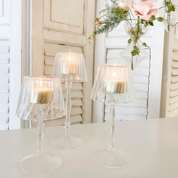 Tre elegante glasfyrfadsstager p� h�je stilke, formet som sm� lamper med t�ndte lys. De er anrettet p� et hvidt bord med hvide tr�-jalousier og h�ngende blomster i baggrunden.