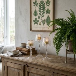 Tre elegante glasfyrfadsstager i forskellige st�rrelser formet som lamper med t�ndte lys, anrettet p� en rustik tr�sk�nk med en gr�n bregne og et botanisk billede p� v�ggen i baggrunden.