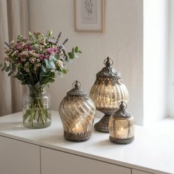 Tre dekorative lanterner i fattigmandsglas med antikt metalm�nster og varmt lys, opstillet p� en hvid sk�nk ved siden af en stor blomsterbuket i en glasvase.
