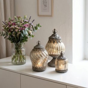 Tre dekorative lanterner i fattigmandsglas med antikt metalm�nster og varmt lys, opstillet p� en hvid sk�nk ved siden af en stor blomsterbuket i en glasvase.