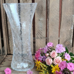En stor, klar glasvase med krakeleret m�nster placeret p� en rustik tr�overflade ved siden af en farverig buket af lyser�de roser, phlox og gule blomster mod en baggrund af r� tr�br�dder.