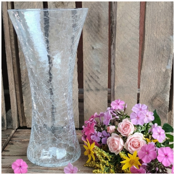 En stor, klar glasvase med krakeleret m�nster placeret p� en rustik tr�overflade ved siden af en farverig buket af lyser�de roser, phlox og gule blomster mod en baggrund af r� tr�br�dder.