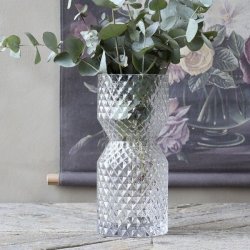 Glasvasen med diamantm�nster brugt til gr�nne eucalyptusgrene placeret p� et rustikt tr�bord foran et blomstermaleri.