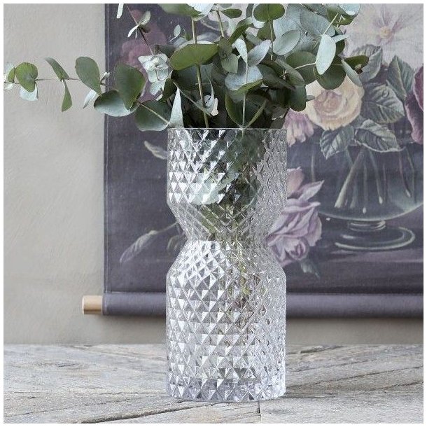 Glasvasen med diamantm�nster brugt til gr�nne eucalyptusgrene placeret p� et rustikt tr�bord foran et blomstermaleri.