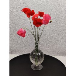 En slank, klar glasvase p� fod med vand og flere r�de og lyser�de valmuer p� lange, gr�nne stilke, placeret p� en sort, rund bakke mod en hvid, m�nstret v�g.