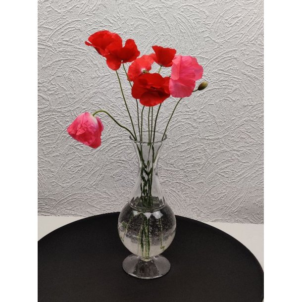 En slank, klar glasvase p� fod med vand og flere r�de og lyser�de valmuer p� lange, gr�nne stilke, placeret p� en sort, rund bakke mod en hvid, m�nstret v�g.
