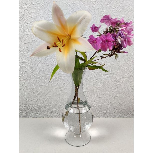 En elegant, timeglasformet glasvase p� fod, fyldt med vand og en smuk buket best�ende af en stor, hvidgylden lilje og klynger af lyser�de blomster, placeret mod en hvid, m�nstret v�g.