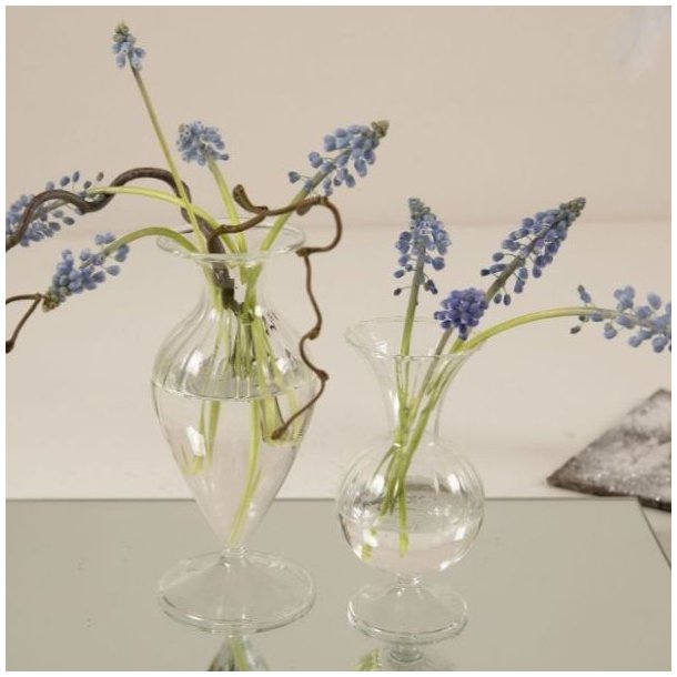 glasvaser-elke-milj�-spejlplade to sm� klare glasvaser med riller st�ende p� en spejlplade p� et d�kket bord med bl� perlehyacinter i.