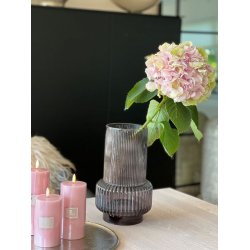 gr�-mila-retro-vase-glas-miljoe Et hyggeligt indend�rs milj�billede, hvor den h�je, riflede retro glasvase i r�ggr� st�r p� et lyst tr�bord med en stor, lyser�d og gr�n hortensiablomst i. Til venstre for vasen ses tre d�mpede lyser�de bloklys med t�ndte LED-flammer st�ende p� en m�rk metalbakke.