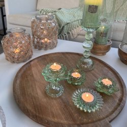 groen-fyrfadsstage-glas-miljoe-samlet-bakke Et stemningsfuldt milj�billede p� en terrasse, hvor fire forskellige assorterede fyrfadsstager i gr�nt glas st�r med t�ndte lys p� en stor rund tr�bakke. I baggrunden ses en lys sofa med gr�nne plaider, store lanterner med rebflet og en h�j m�nstret glasstage med et gr�nt bloklys.