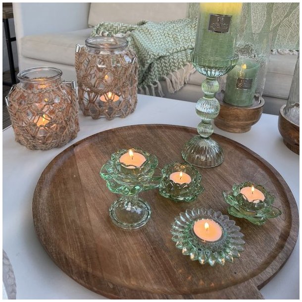 groen-fyrfadsstage-glas-miljoe-samlet-bakke Et stemningsfuldt milj�billede p� en terrasse, hvor fire forskellige assorterede fyrfadsstager i gr�nt glas st�r med t�ndte lys p� en stor rund tr�bakke. I baggrunden ses en lys sofa med gr�nne plaider, store lanterner med rebflet og en h�j m�nstret glasstage med et gr�nt bloklys.