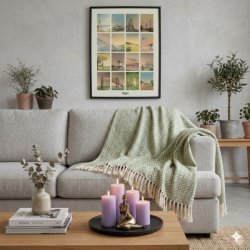 Et firkantet milj�billede af en gr�n plaid med hvidt sildebensm�nster og dekorative frynser, der er kastet henover rygl�net p� en lys gr� sofa. P� sofabordet i forgrunden st�r et sort fad med lilla bloklys og en lille bronzefigur, mens baggrunden prydes af gr�nne planter og et indrammet kunstv�rk p� v�ggen, hvilket skaber en hyggelig og moderne stueatmosf�re.