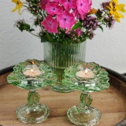 To gr�nne blomsterformede fyrfadsstager i glas p� stilk med t�ndte lys foran en gr�n rillet vase med en farverig buket blomster.