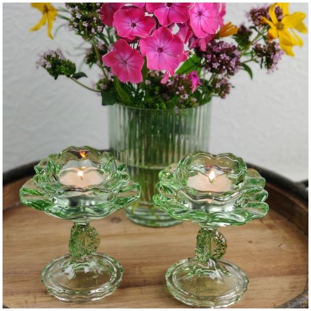 To gr�nne blomsterformede fyrfadsstager i glas p� stilk med t�ndte lys foran en gr�n rillet vase med en farverig buket blomster.