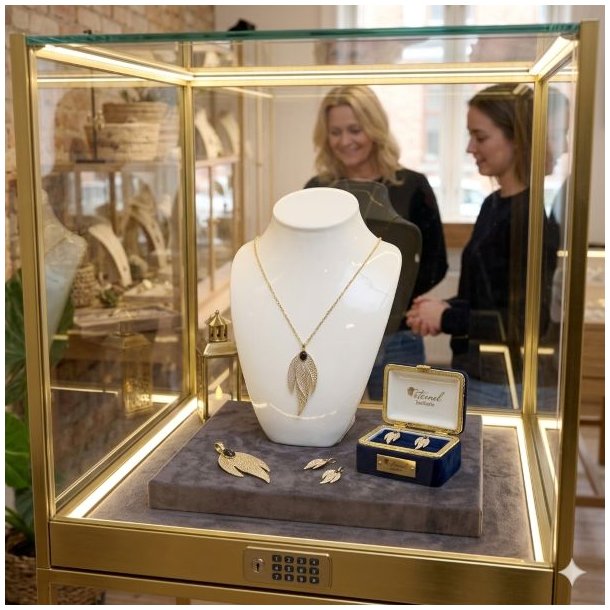 En forgyldt La Vida guldhalsk�de med trebladet vedh�ng og sort sten udstillet p� en hvid porcel�nsholder i en oplyst glasmontre.