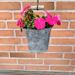 Et n�rbillede af en rustik, patineret h�ngende zinkurtepotte-spand, der er fyldt med livlige, pink flittiglise-blomster. Spanden har en metalt hank med et distinkt, lyst tr�greb og h�nger fra en jernstang mod en r�d murstensv�g som en del af en havedekoration.