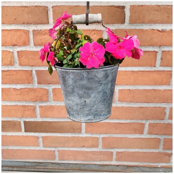 Et n�rbillede af en rustik, patineret h�ngende zinkurtepotte-spand, der er fyldt med livlige, pink flittiglise-blomster. Spanden har en metalt hank med et distinkt, lyst tr�greb og h�nger fra en jernstang mod en r�d murstensv�g som en del af en havedekoration.