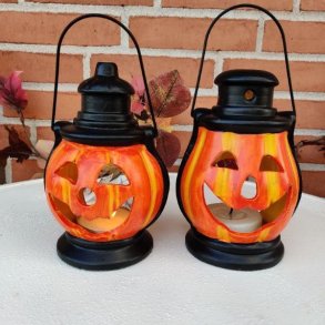 To hyggelige, orange halloween-gr�skarlanterner i keramik med t�ndte fyrfadslys indeni, st�ende p� et hvidt havebord.