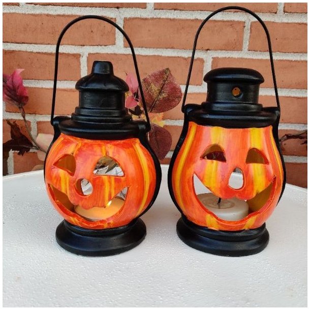 To hyggelige, orange halloween-gr�skarlanterner i keramik med t�ndte fyrfadslys indeni, st�ende p� et hvidt havebord.