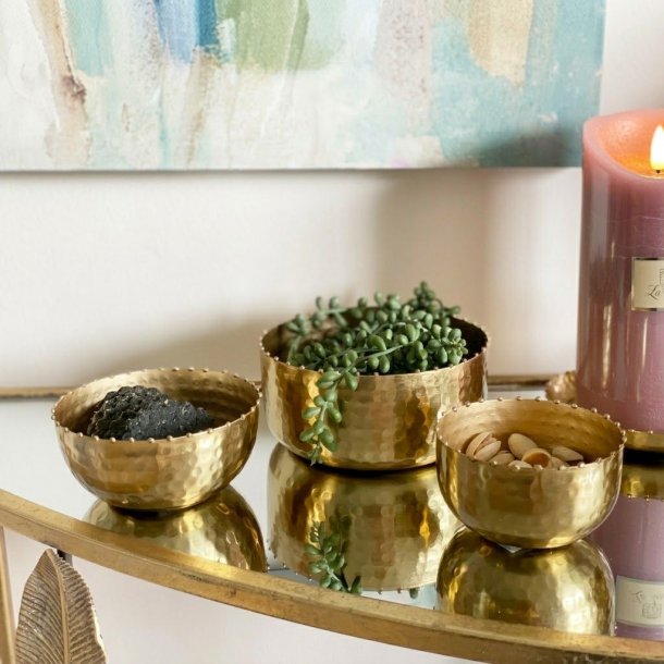 Kvadratisk milj�billede, der viser tre hamrede guld metalsk�le med n�dder, sorte sten og sukkulent planter, placeret p� et spejlbord. Der er et stort lilla bloklys med guld-emblem ved siden af.