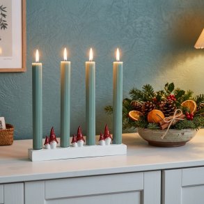 Stemningsfuldt firkantet milj�billede af Harvesttime adventsstagen med t�ndte gr�nne lys placeret p� en hvid sk�nk ved siden af en juledekoration.