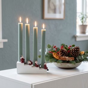 Stemningsfuldt firkantet milj�billede af Harvesttime adventsstagen med t�ndte gr�nne lys placeret p� en hvid sk�nk ved siden af en juledekoration.