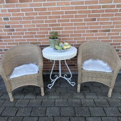 To naturfarvede flettede havestole placeret ved et bord, hvor den forreste stol er stylet med et firkantet hvidt imiteret lammeskind som s�dehynde.