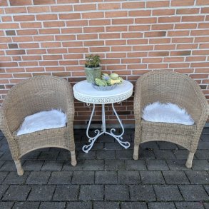 To naturfarvede flettede havestole placeret ved et bord, hvor den forreste stol er stylet med et firkantet hvidt imiteret lammeskind som s�dehynde.